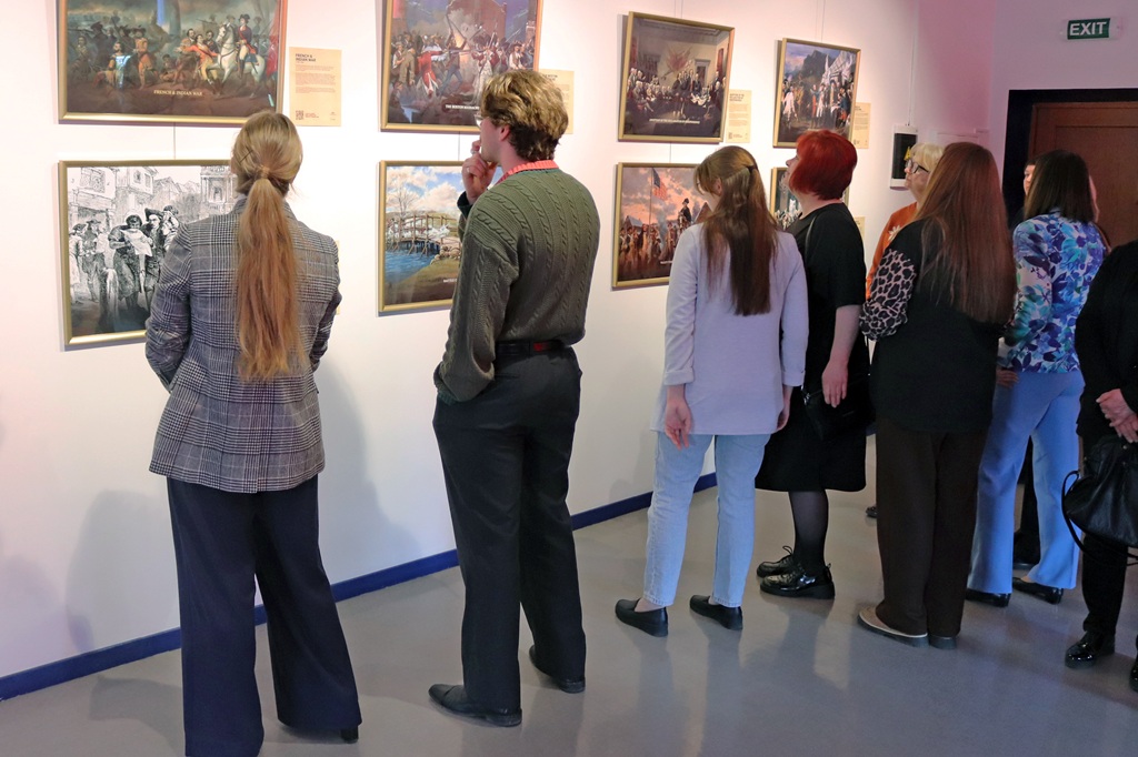 Galerijā cilvēki uzmanīgi vēro gleznas uz baltas sienas. Telpā valda mierīga atmosfēra, un apmeklētāji šķiet dziļi koncentrējušies.