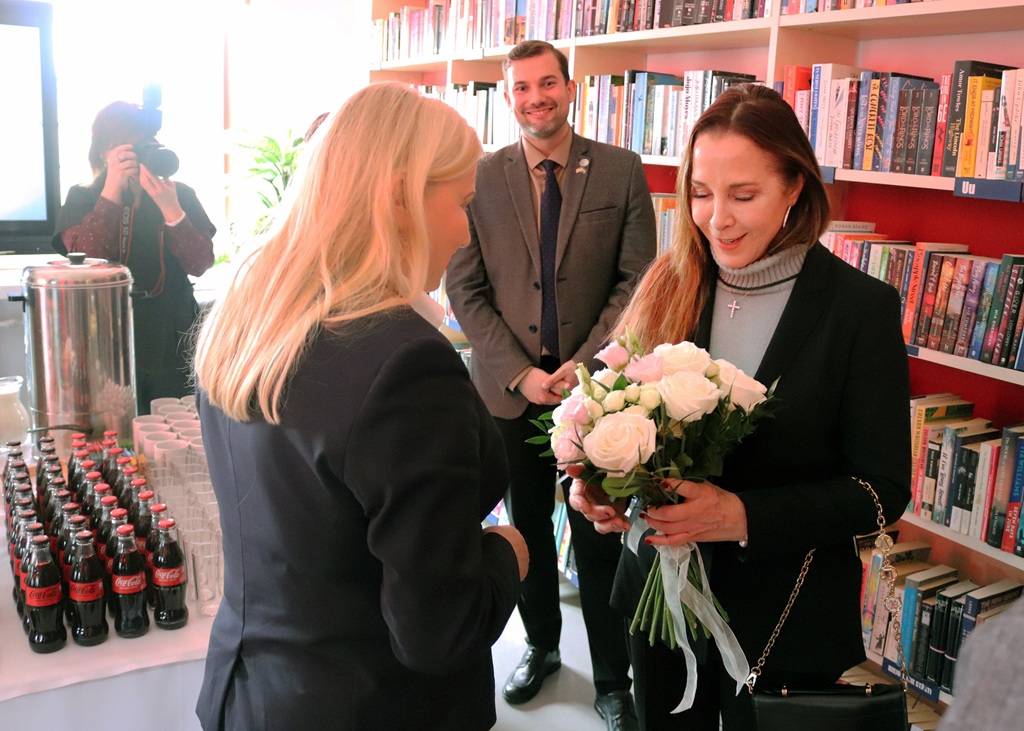 Sieviete melnā mētelī saņem baltu rožu pušķi no citas sievietes ar blondiem matiem. Viņas atrodas grāmatnīcā. Netālu smaida vīrietis uzvalkā. Kreisajā pusē uz galda stāv pudeles un krūzes, un fotogrāfs iemūžina šo mirkli.