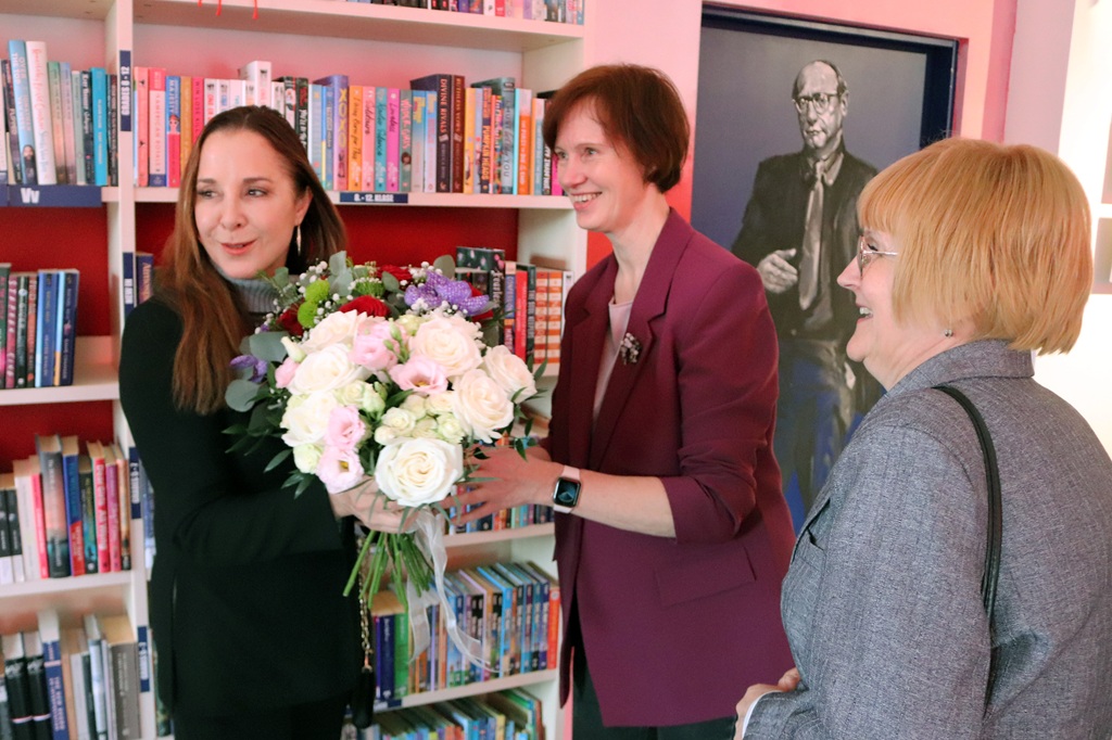 Trīs sievietes stāv bibliotēkā. Viena sieviete melnā tērpā priecīgi tur ziedu pušķi. Otra sieviete bordo krāsas žaketē pasniedz viņai pušķi, silti smaidot. Netālu stāv trešā sieviete, arī smaidot. Fonā grāmatu plaukti ir pilni ar krāsainām grāmatām, un tur ir glezna, kurā attēlots vīrietis. Atmosfēra ir draudzīga un svinīga.