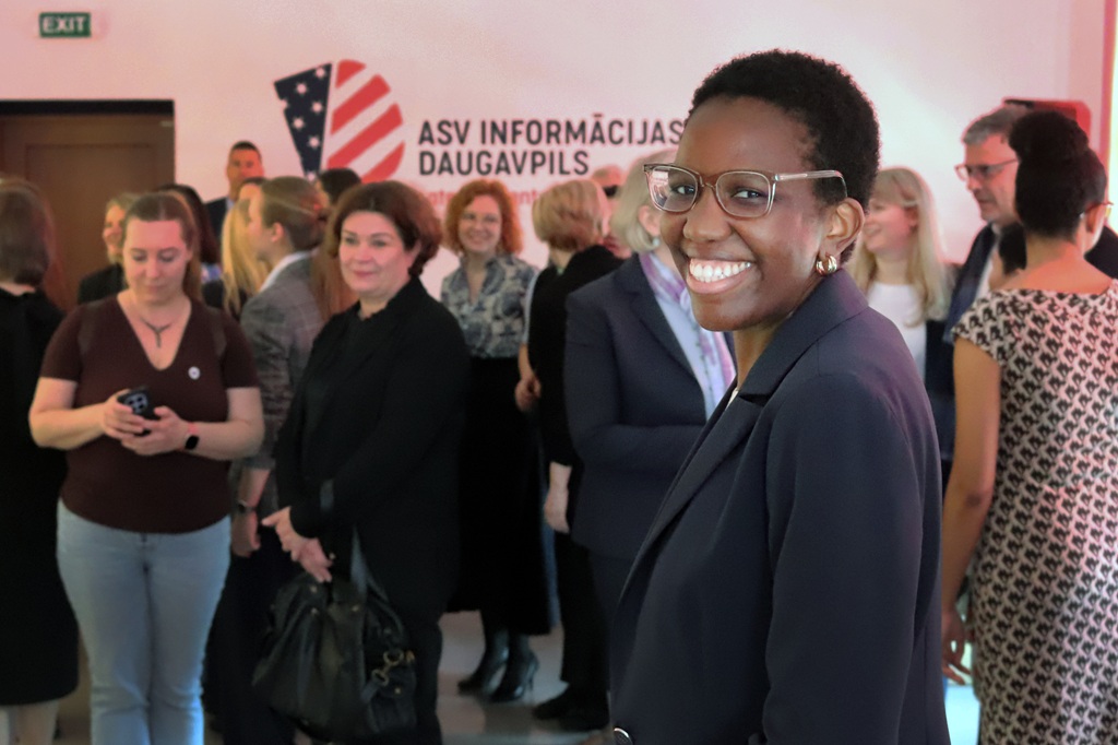 Cilvēks brillēs un tumšā žaketē smaida kamerai rosīgā telpā. Fonā rosās dažādu cilvēku grupa, radot dzīvīgu atmosfēru.