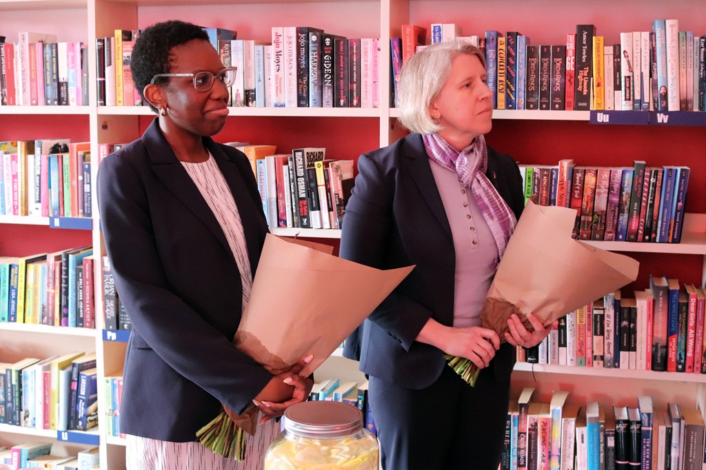 Divi cilvēki stāv bibliotēkā, turot rokās brūnā papīrā ietītus pušķus. Viņi ir ģērbušies svinīgi, ieskauti krāsainos grāmatu plauktos un izskatās domīgi.