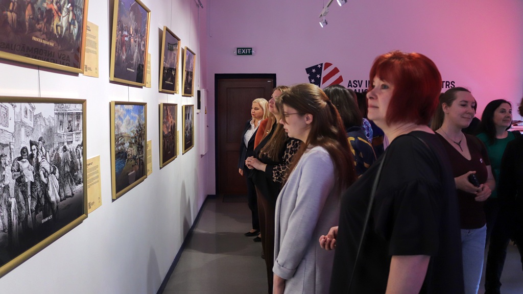 Sieviešu grupa uzmanīgi aplūko ierāmētus mākslas darbus uz galerijas sienas. Telpa ir maigi apgaismota, radot pārdomu rosinošu un saistošu atmosfēru.