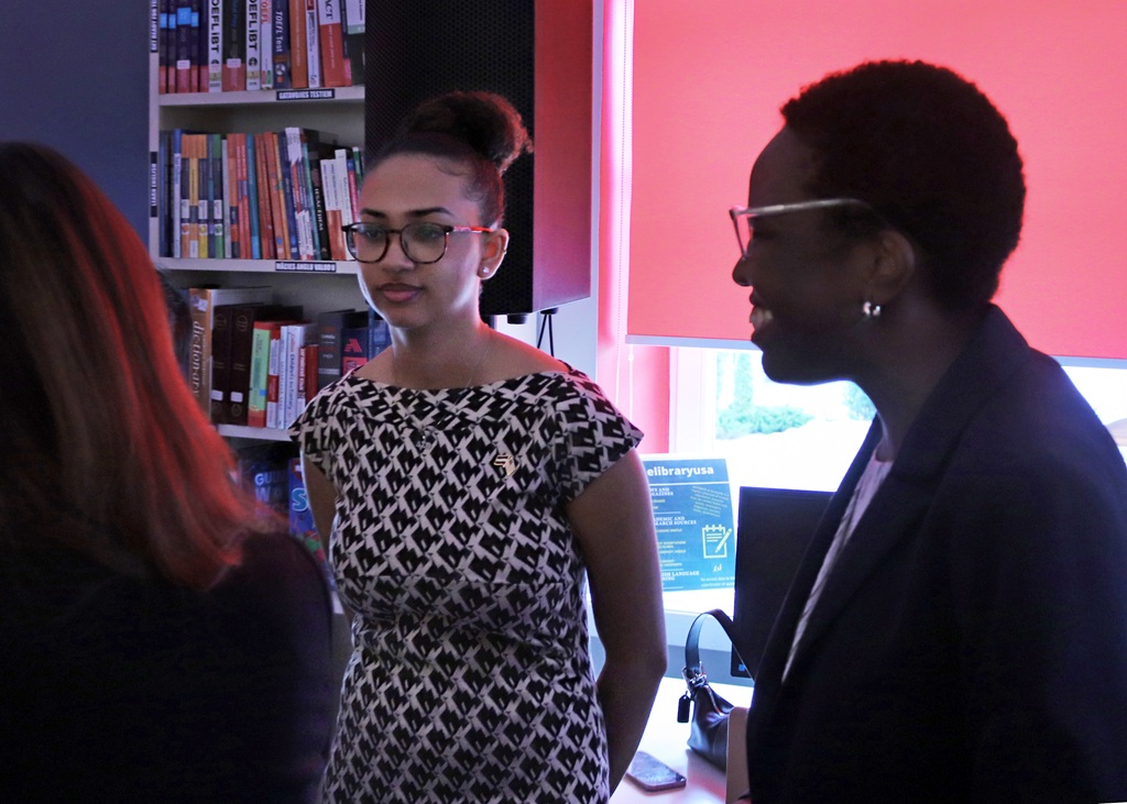 Divas sievietes sarunājas bibliotēkā, aiz kurām atrodas grāmatu plaukti. Viena valkā brilles un rakstainu kleitu, otra smaida žaketē. Silts apgaismojums rada draudzīgu atmosfēru.