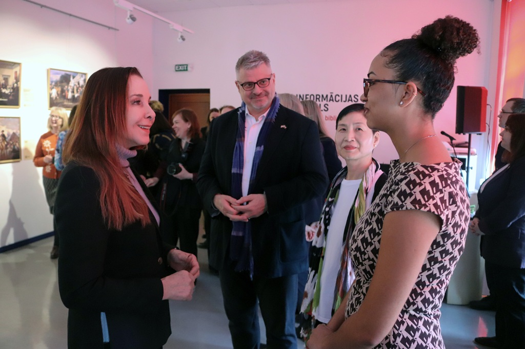 Četru cilvēku grupa labi apgaismotā galerijā iesaistās sarunā, ko ieskauj sienas mākslas darbi. Noskaņojums ir draudzīgs un profesionāls.