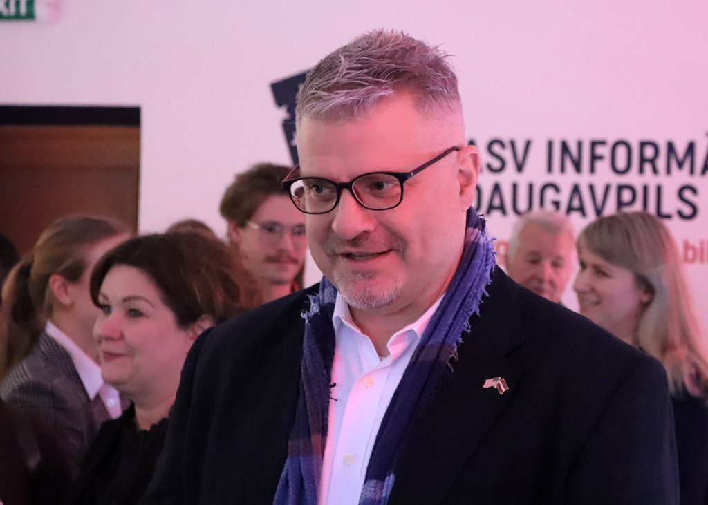 Vīrietis ar brillēm un šalli smaida pasākumā. Viņš stāv izplūdušas grupas priekšā, fonā daļēji redzama zīme.