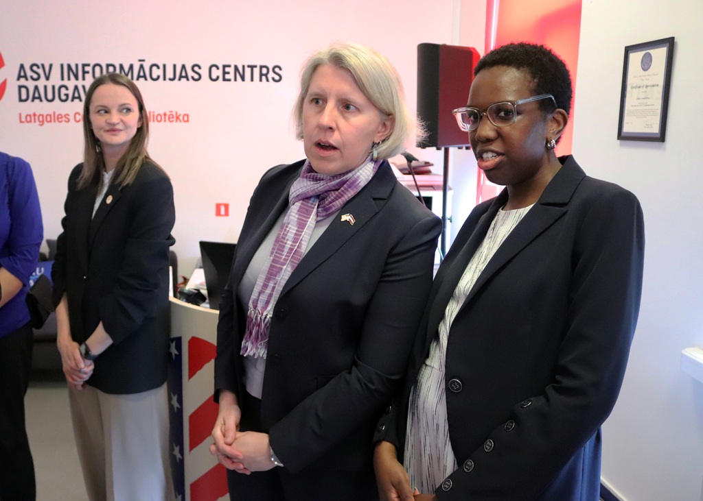 Trīs sievietes lietišķā apģērbā stāv telpā, pie kuras sienas ir uzraksts "ASV Informācijas Centrs". Viņas šķiet iesaistītas un koncentrētas. Atmosfēra ir profesionāla.