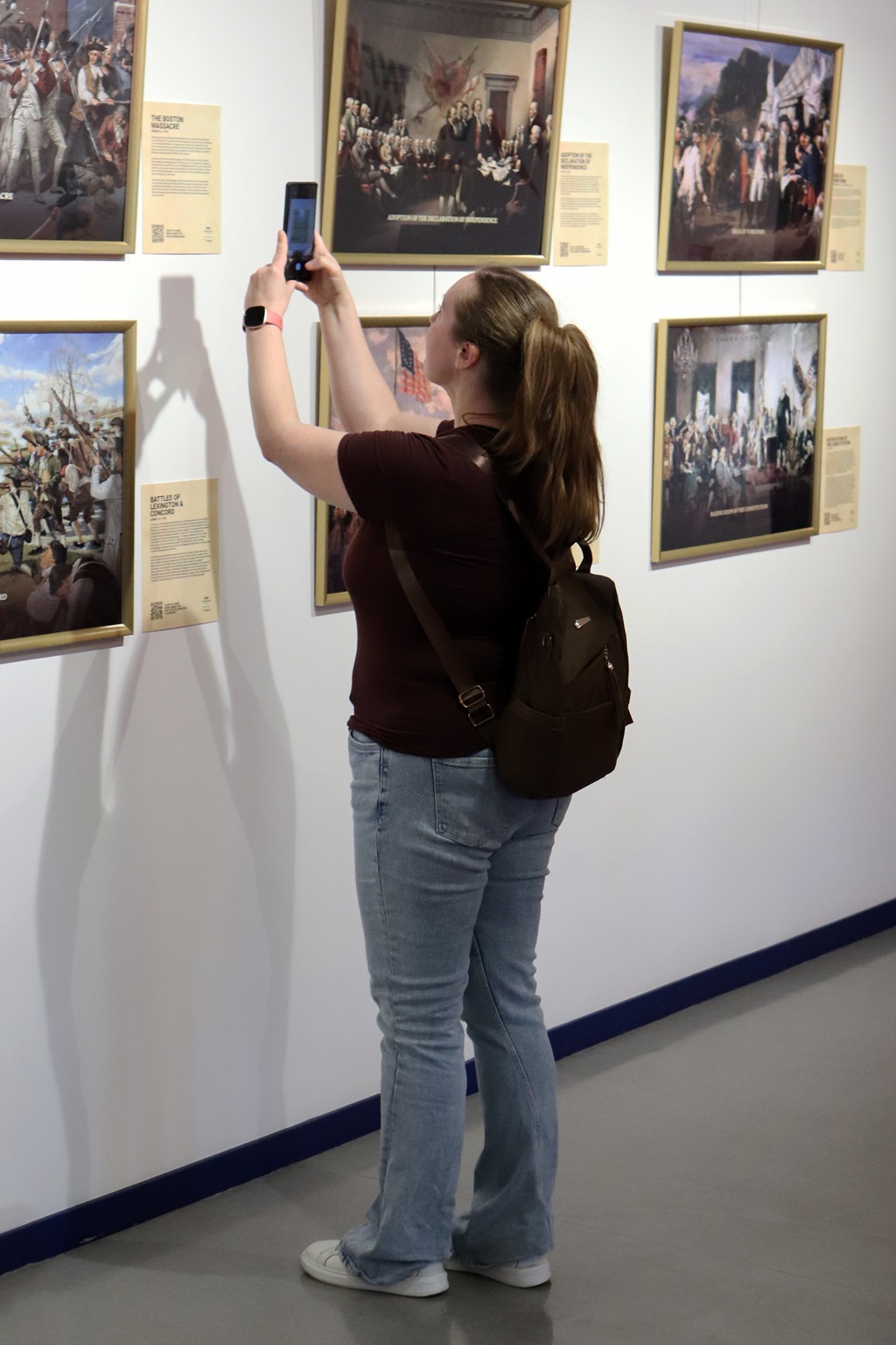 Sieviete ar zirgasti fotografē gleznas galerijā. Viņai mugurā ir džinsi, sarkanbrūns krekls un mugursoma. Aina ir mierīga un koncentrēta.