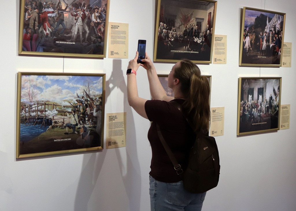 Sieviete fotografē galerijas sienu, kurā ir ierāmētas vēsturiskas gleznas. Aina pauž interesi un izglītojošu iespaidu, ko ieskauj informatīvas plāksnes.