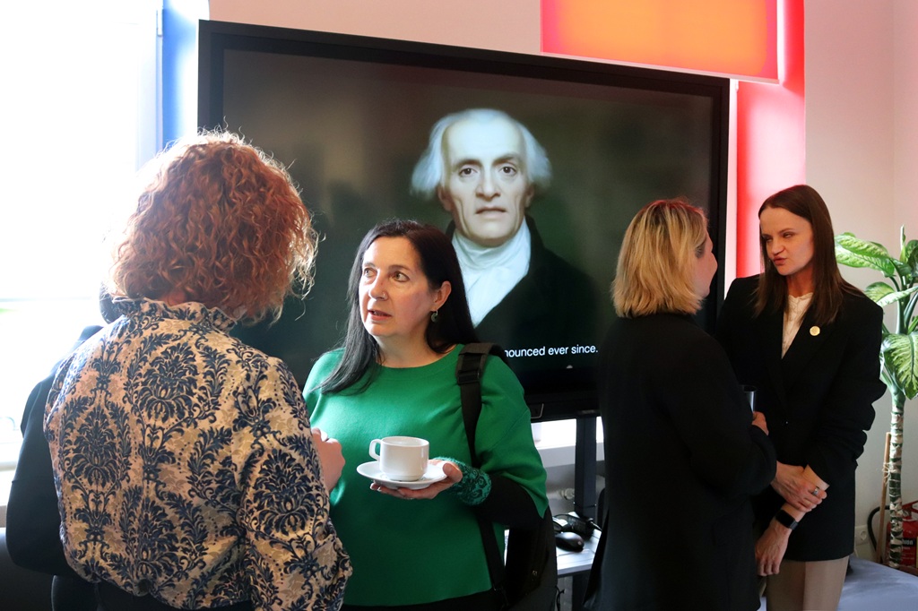 Četras sievietes sarunājas modernā telpā. Aiz viņām uz ekrāna ir attēlots portrets. Atmosfēra ir profesionāla un sabiedriska.