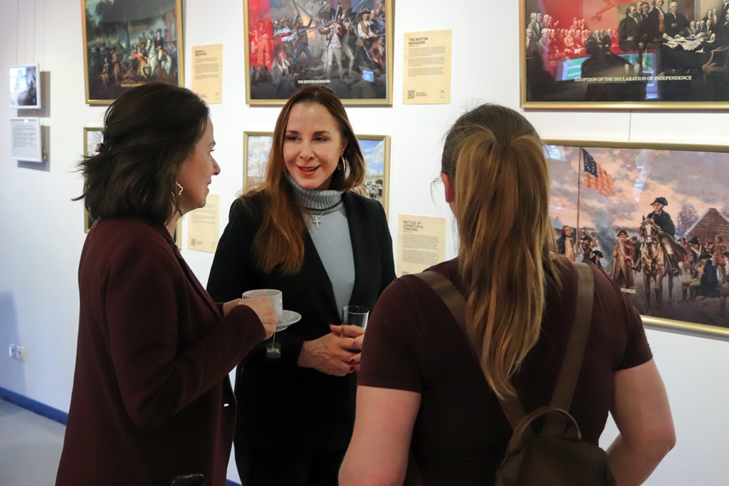 Trīs cilvēki sarunājas mākslas izstādē. Viņi stāv smaidoši, ieskauti vēsturiskām gleznām uz baltas sienas, radot siltu atmosfēru.