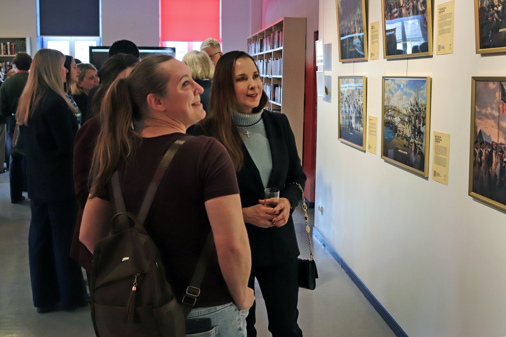 Divas sievietes mākslas galerijā apbrīno ierāmētas fotogrāfijas uz baltas sienas. Viena tur dzērienu, abas smaida, apkārt ir citi apmeklētāji. Silta, valdzinoša atmosfēra.