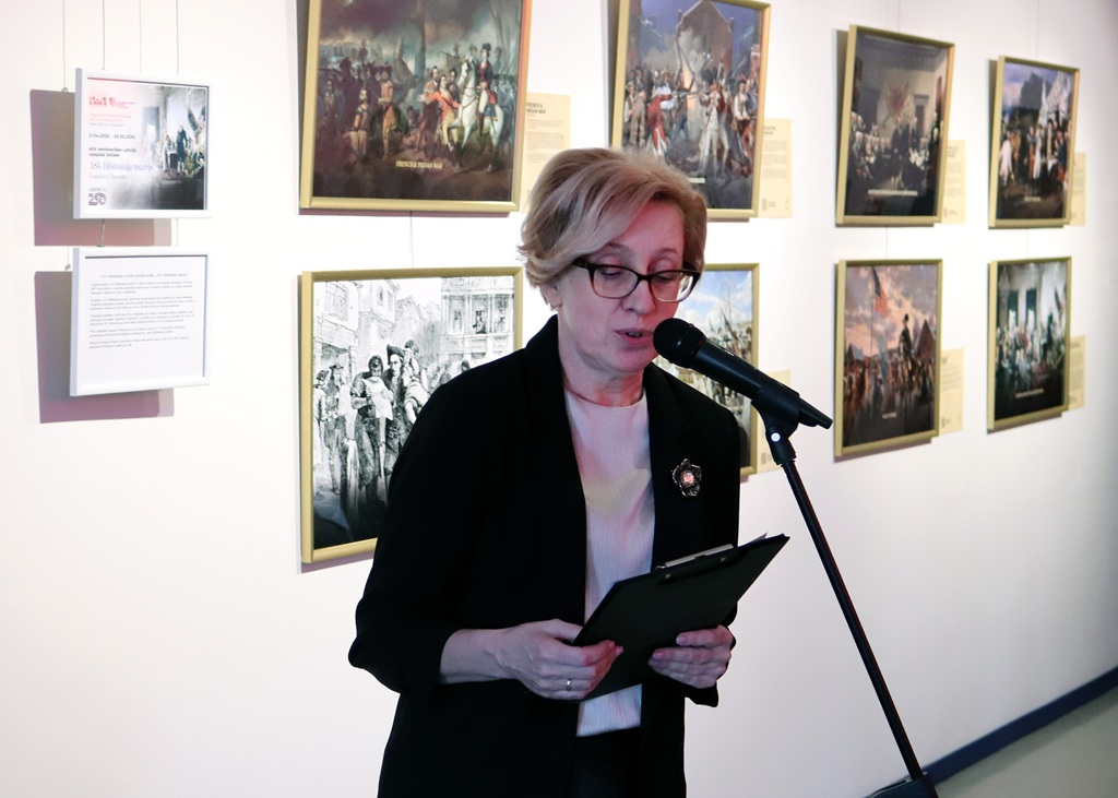 Sieviete runā mikrofonā. Viņa tur rokās planšeti un stāv vairāku ierāmētu gleznu priekšā pie sienas, radot formālu un saistošu atmosfēru.