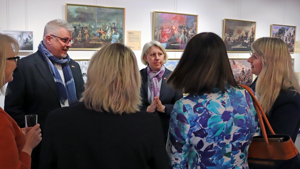 Sešu cilvēku grupa sarunājas mākslas galerijā, pie sienām karājoties krāsainām gleznām. Atmosfēra ir sabiedriska un rosīga.