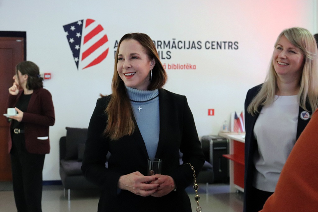 Divas sievietes smaida profesionālā vidē ar amerikāņu tematikas logotipu pie sienas. Viena tur glāzi, bet otra fonā tur krūzi.