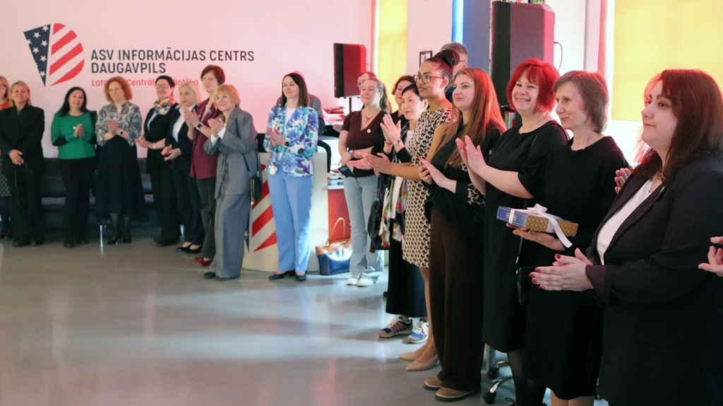 Cilvēku grupa stāv puslokā telpās, smaida un aplaudē. Fonā ir redzama zīme, kas norāda ASV Informācijas centru Daugavpilī.