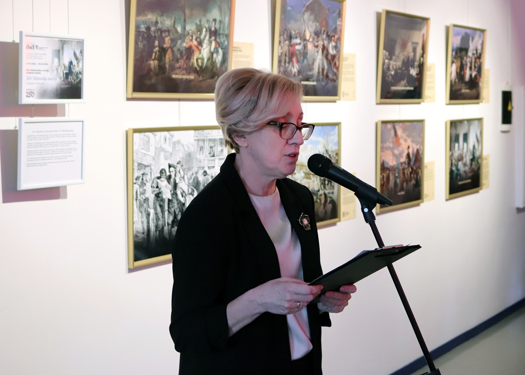 Sieviete brillēs runā mikrofonā, turot rokās planšetdatoru mākslas galerijā. Aiz viņas pie sienas rotā gleznas zelta rāmjos.