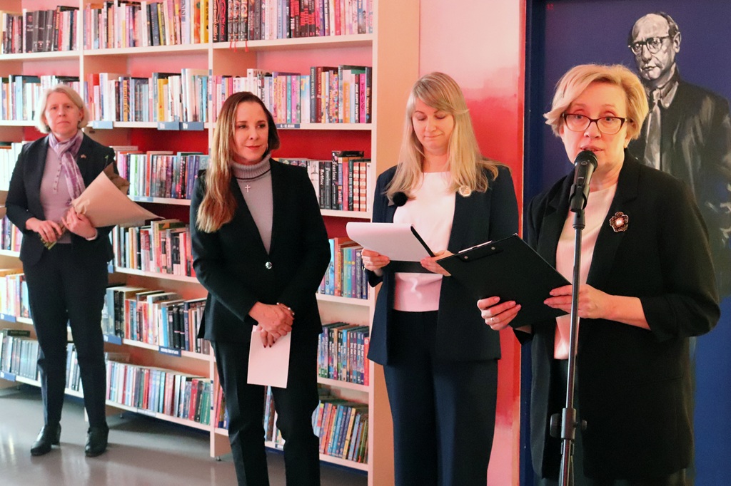 Sieviete runā mikrofonā bibliotēkas vidē, turot rokās planšeti. Blakus viņai stāv vēl trīs sievietes, viena tur ziedus, visas izskatās formālas.