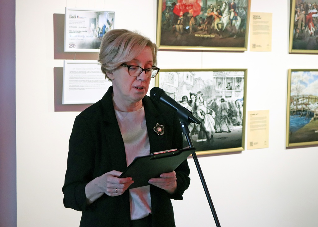 Sieviete ar īsiem blondiem matiem un brillēm runā mikrofonā, turot rokās planšetdatoru. Viņa stāv ierāmēta mākslas darba priekšā uz galerijas sienas.