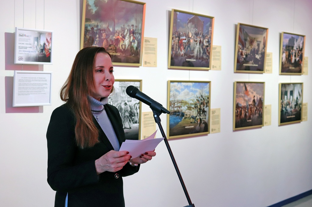 Sieviete runā mikrofonā, turot rokās piezīmes. Aiz viņas uz baltas sienas ir izliktas vairākas ierāmētas gleznas.