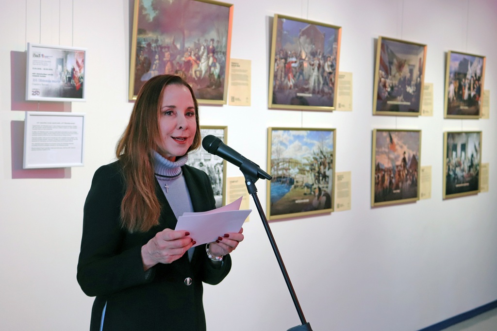 Sieviete, turot papīrus, runā mikrofonā. Aiz viņas pie sienas karājas ierāmētas gleznas, radot kulturālu atmosfēru.