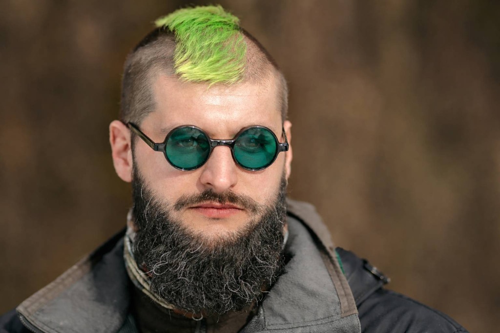 Vīrietis ar spilgti zaļu mohauku un kuplu bārdu valkā apaļas, zaļganas brilles. Viņš izskatās mierīgs, ģērbies daudzslāņainās jakās uz izplūduša brūna fona.