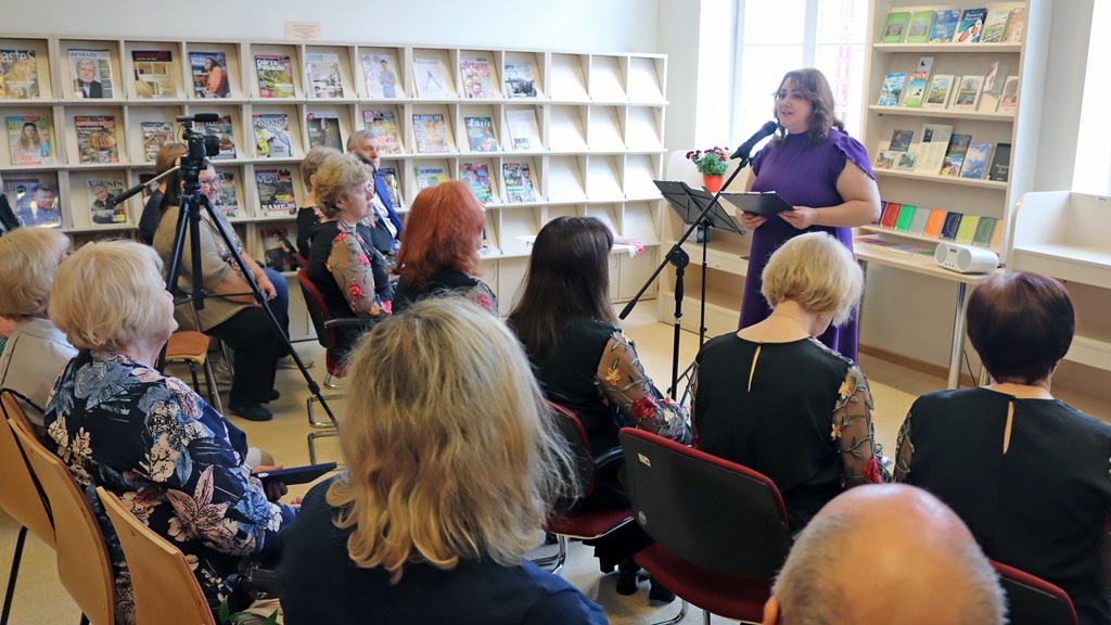 Sieviete purpursarkanā kleitā uzstājas no tribīnes labi apgaismotā telpā, uzrunājot uzmanīgu auditoriju. Gar sienām stāv plaukti ar žurnāliem un grāmatām.