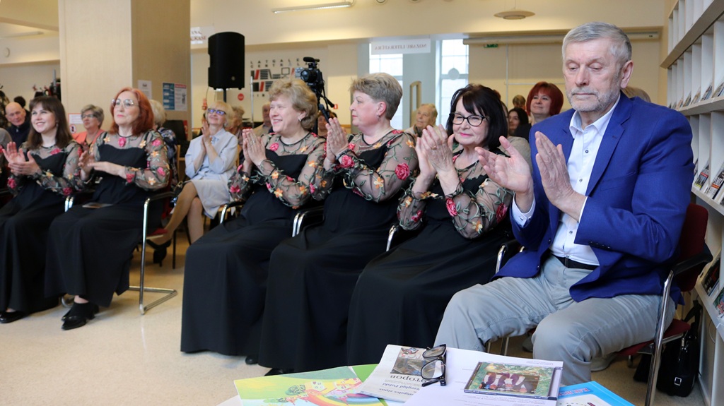 Cilvēki sēž gaišā telpā un aplaudē. Sievietes ziedu raksta kleitās un vīrietis zilā žaketē šķiet ieinteresēti un pateicīgi. Uz galda papīri un grāmatas.