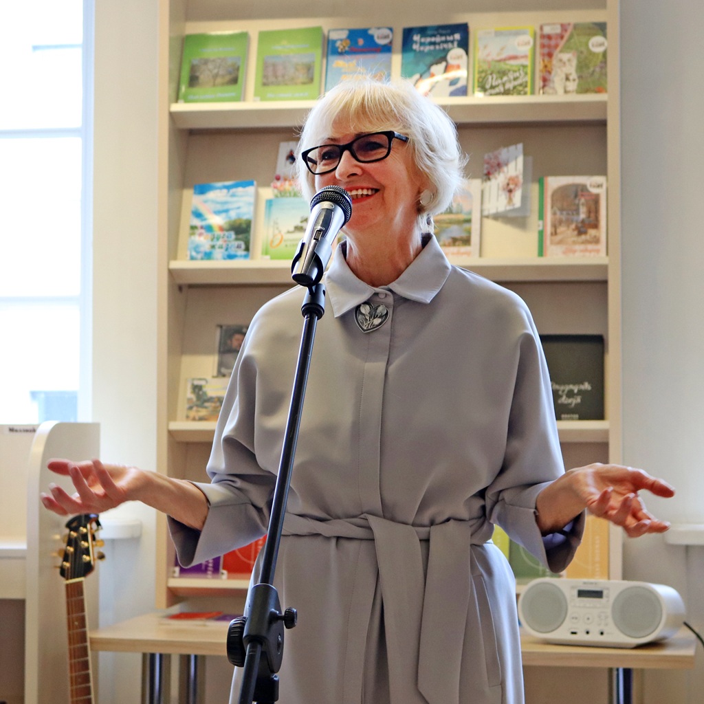 Vecāka gadagājuma sieviete ar sirmiem matiem un brillēm entuziastiski runā mikrofonā. Viņa ir ģērbusies gaiši pelēkā kleitā un stāv mājīgā grāmatnīcā.