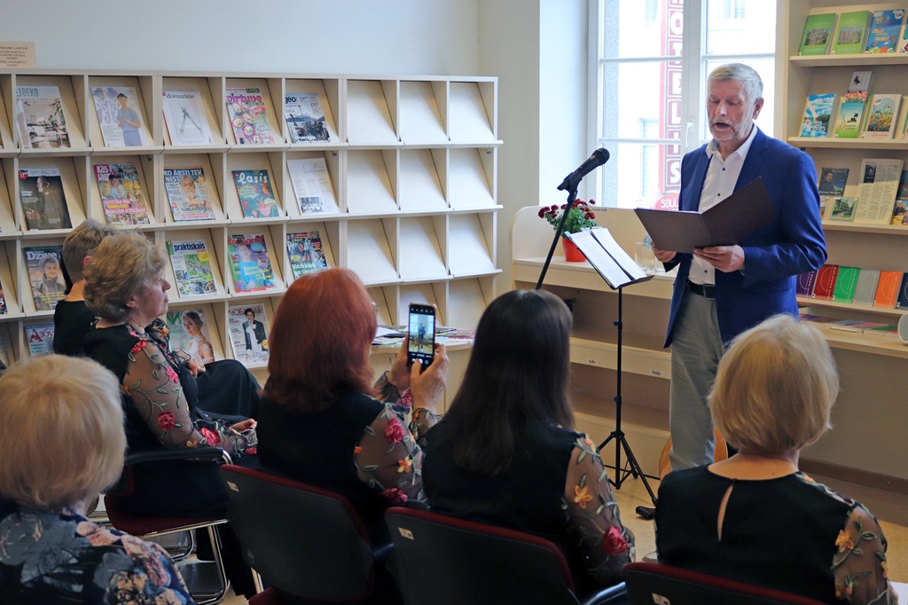 Vīrietis stāv pie mikrofona, lasot no mapes uzmanīgai auditorijai bibliotēkas vidē. Plauktos fonā ir izvietotas grāmatas un žurnāli. Sieviete auditorijā fotografē ar savu tālruni, radot dzīvīgu un saistošu atmosfēru.