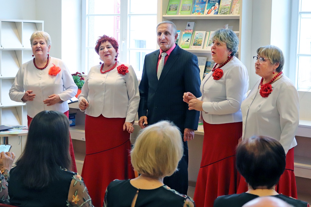 Četru sieviešu un viena vīrieša grupa uzstājas telpās. Sievietes ģērbušās vienādos baltos topos un sarkanos svārkos ar ziedu rakstiem. Skatītāji uzmanīgi vēro.