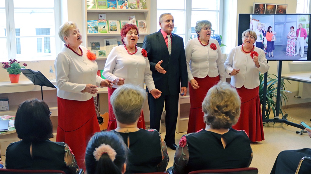 Četru sieviešu un viena vīrieša grupa, ģērbušies saskaņotā sarkanā un baltā apģērbā, kaislīgi dzied auditorijas priekšā gaišā, ar grāmatām piepildītā telpā.