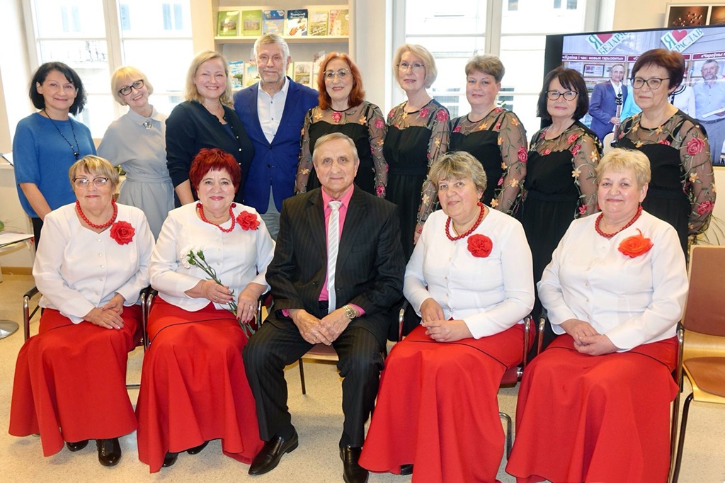 Grupa gados vecāku sieviešu un vīriešu pozē telpās, silti smaidot. Sievietes vienādos baltos blūzēs, sarkanos svārkos un sarkanos ziedos. Vīrieši uzvalkos. Aiz viņiem grāmatu plaukti.