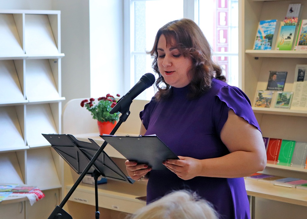 Sieviete purpursarkanā kleitā runā mikrofonā, turot rokās mapi. Viņa atrodas bibliotēkā, kur redzamas grāmatas un podā iestādīts augs, radot koncentrētu un iesaistītu atmosfēru.