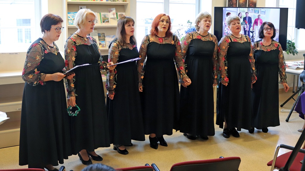 Septiņas sievietes dzied korī, tērptas vienādos melnos kleitās ar ziedu raksta piedurknēm. Viņas izskatās koncentrētas un izteiksmīgas, radot siltu, harmonisku sajūtu.