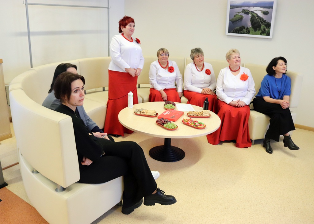 Septiņas sievietes sēž pusapļa bēšā dīvānā, ģērbušās baltos kreklos ar sarkaniem svārkiem. Uz galda atrodas uzkodas. Aina šķiet mierīga un kopīga.