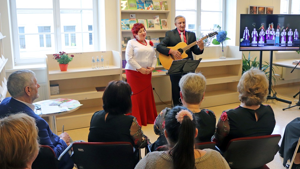 Vīrietis spēlē ģitāru, kamēr sieviete dzied auditorijai gaišā telpā ar grāmatu plauktiem. Ekrānā redzami izpildītāji tradicionālā apģērbā, piešķirot kultūras pieskaņu.