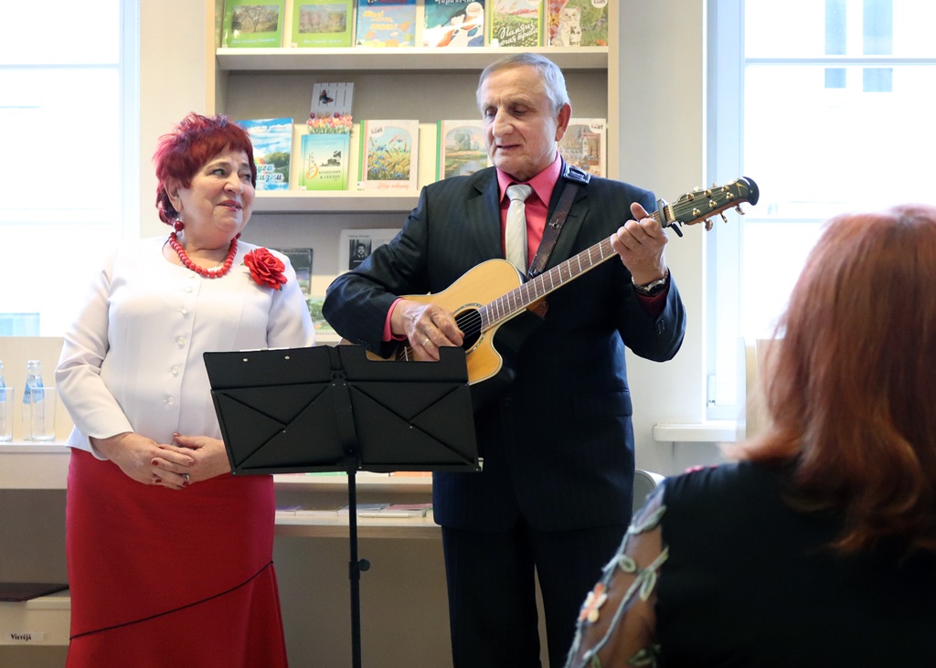 Vecāks vīrietis uzvalkā spēlē ģitāru blakus vecākai sievietei baltā blūzē un sarkanā svārkos, kura smaida. Viņi atrodas gaišā istabā, kuras plauktos ir sakrautas grāmatas.