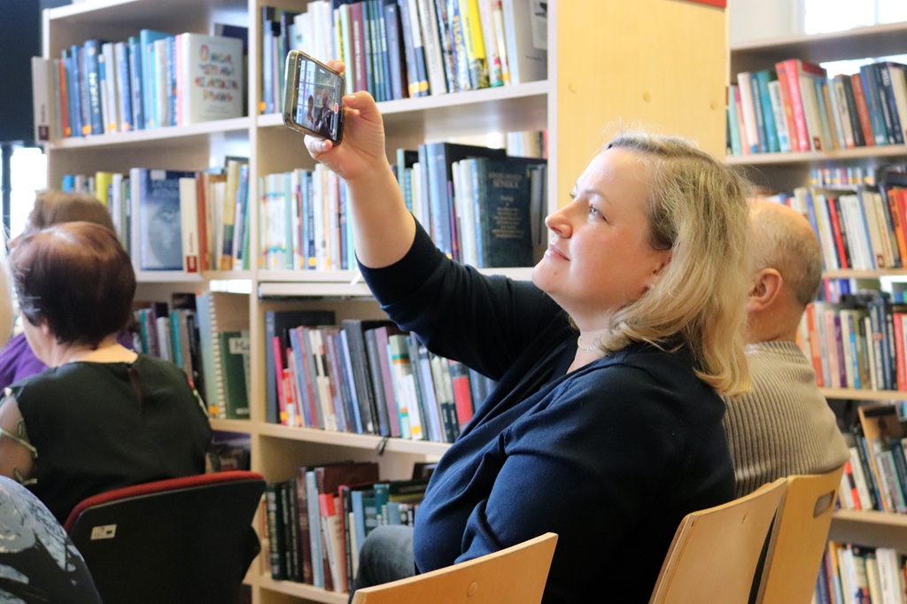 Sieviete bibliotēkā uzņem selfiju ar savu viedtālruni, ko ieskauj grāmatu plaukti. Viņa izskatās apmierināta un koncentrēta, sēžot starp citiem.