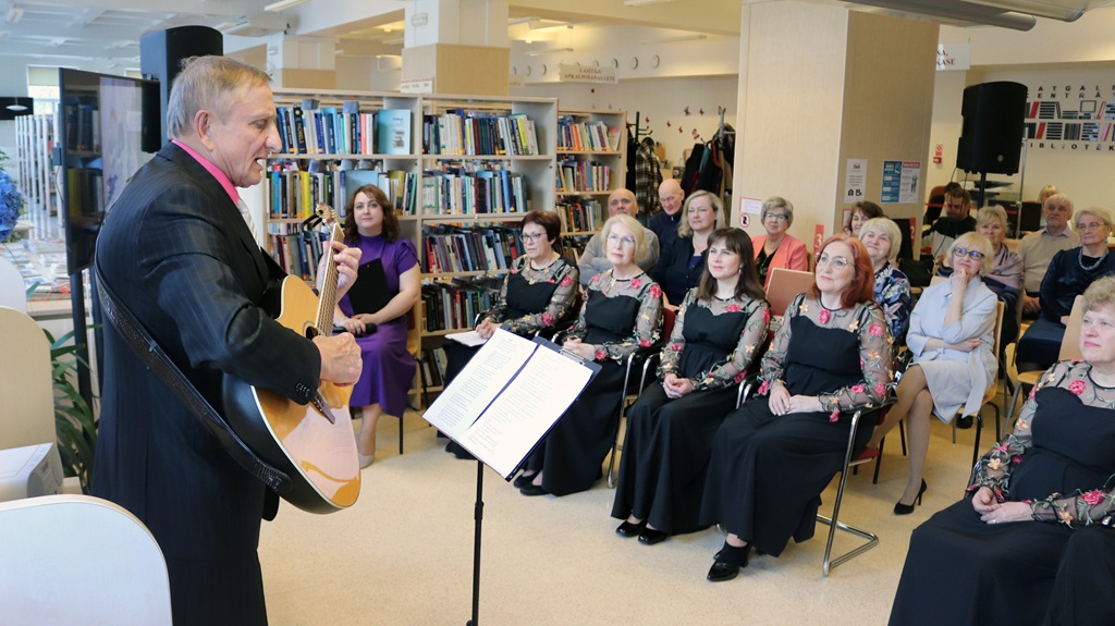 Vīrietis uzvalkā spēlē ģitāru uzmanīgai auditorijai, kas sēž bibliotēkā. Sievietes vienādos tērpos sēž pirmajā rindā, radot mājīgu un saistošu atmosfēru.