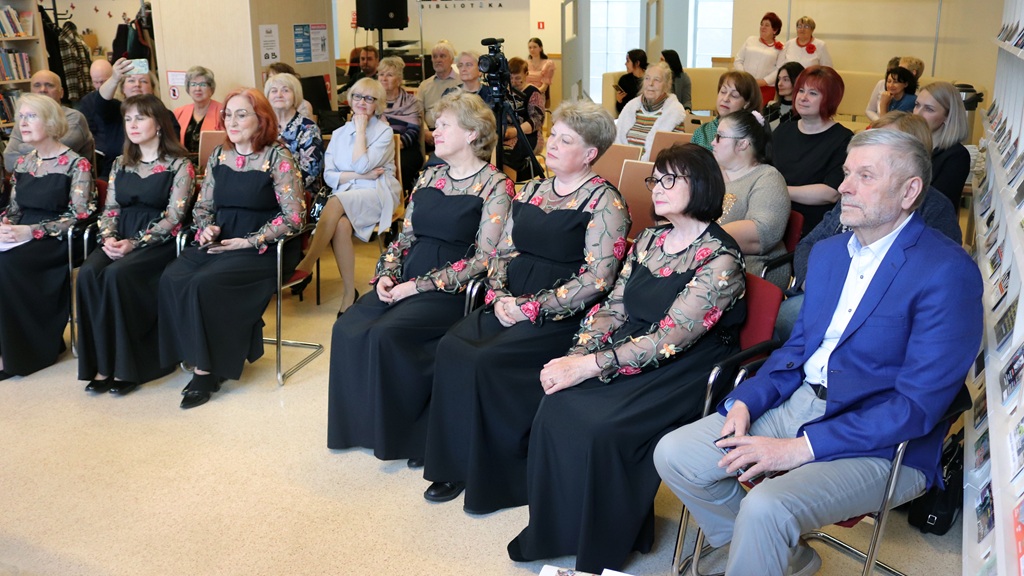 Auditorija uzmanīgi sēž, redzamas sievietes ziedu raksta kleitās un vīrietis zilā žaketē. Vide atgādina pasākumu vai prezentāciju telpās.