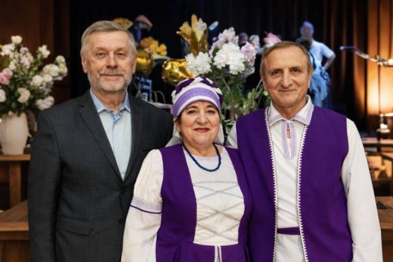 Trīs cilvēki stāv ziedu kompozīcijas priekšā. Divi ir ģērbušies purpursarkanos un baltos tautastērpos, bet otrs ir uzvalkā un sirsnīgi smaida.