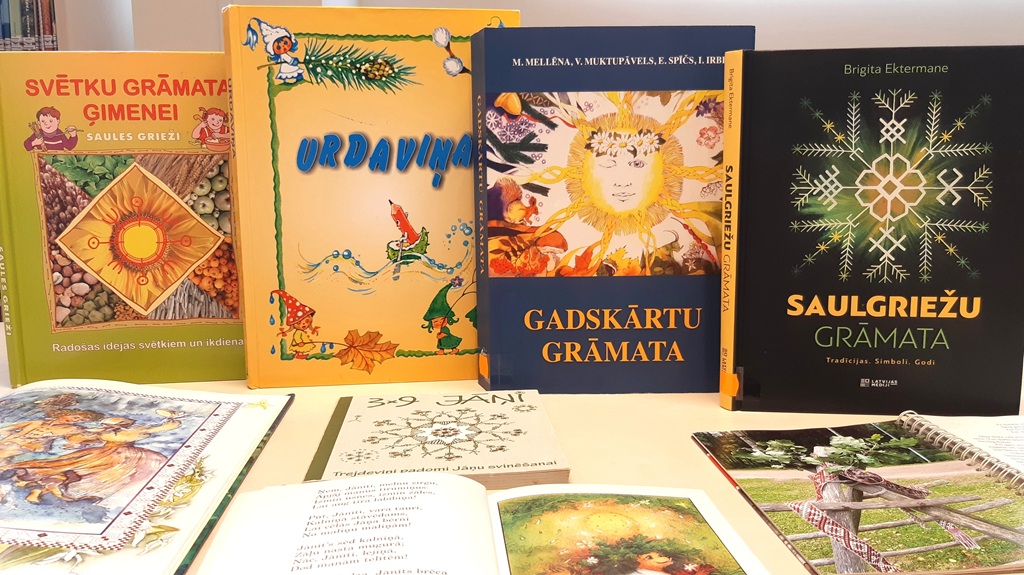 Četras krāsainas grāmatas izkārtotas vertikāli, atspoguļojot kultūras tēmas - Jāņi, saulgrieži, gadskārtas, svētku grāmata ģimenei. Priekšplānā novietotas atvērtas grāmatas.
