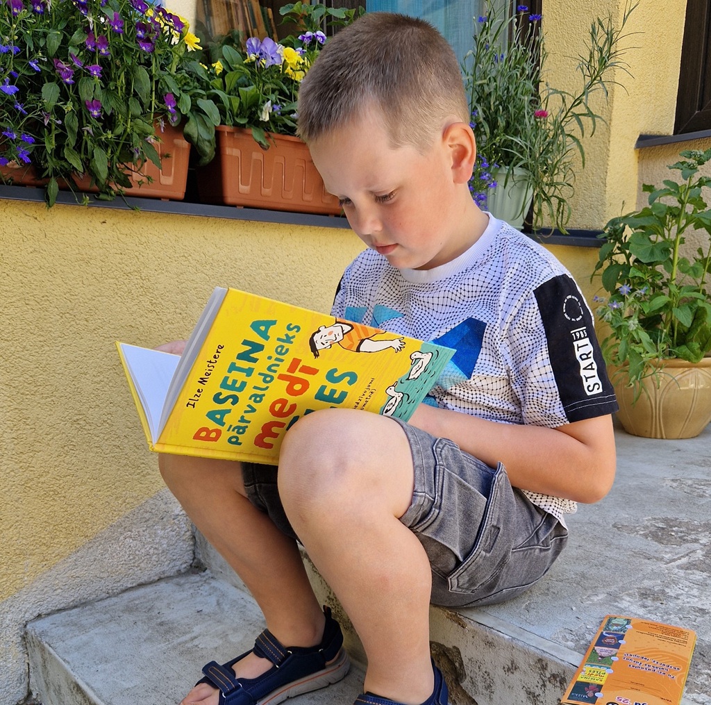Mazs zēns, kurš sēž uz betona pakāpieniem, lasa krāsainu grāmatu. Viņam mugurā ir krekliņš ar uzrakstiem, šorti un sandeles. Viņam aizmugurē podos zied košas puķes. Vasarīgas noskaņas.