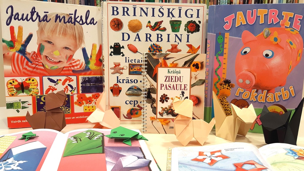 Uz galda blakus paštaisītiem origami dzīvniekiem izvietotas krāsainas rokdarbu grāmatas latviešu valodā. Skats pauž radošumu un rotaļīgu izpēti.