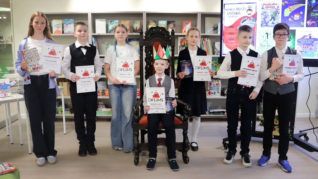 Septiņi bērni stāv bibliotēkā, lepni turot rokās sertifikātus, viens bērns sēž lielā, greznā krēslā ar papīra kroni galvā. Noskaņojums ir svinīgs.