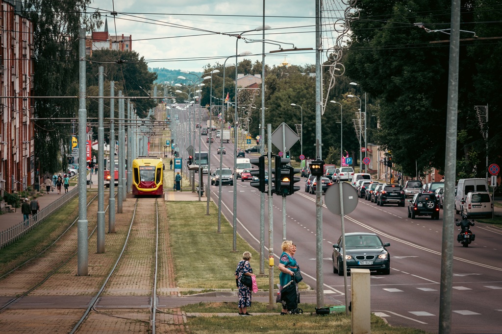 Pilsētas ielas aina ar tramvaja sliedēm kreisajā pusē, tuvojas sarkans un dzeltens tramvajs. Cilvēki gaida pie pārbrauktuves, automašīnas brauc pa ceļu. Koki un ēkas ieskauj ielu.