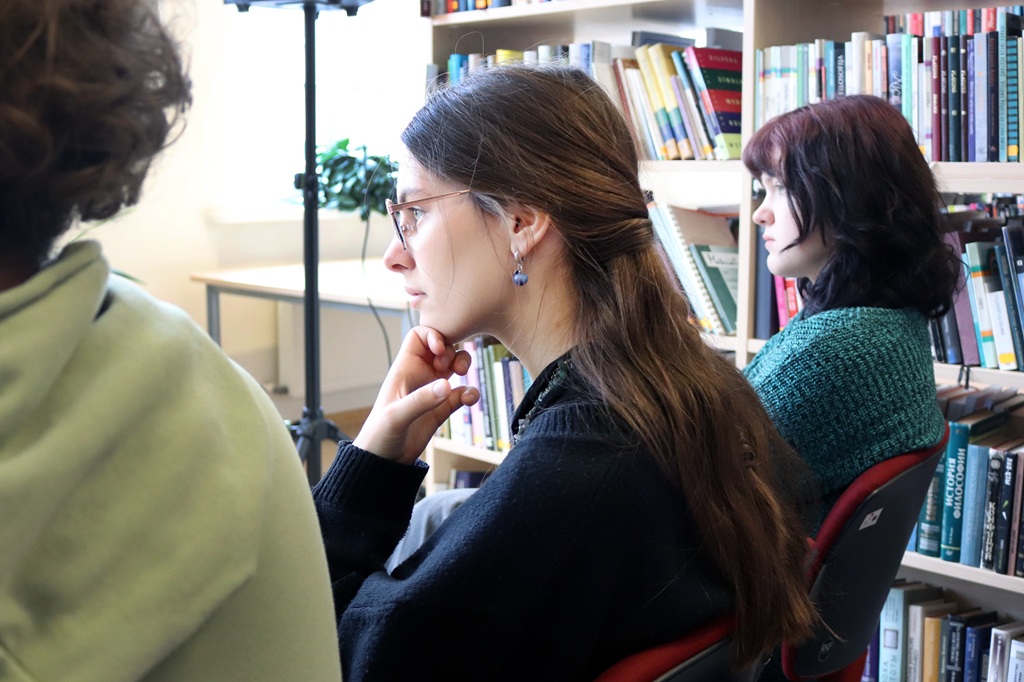 Divi cilvēki uzmanīgi klausās bibliotēkas sesijas laikā. Sieviete brillēs ar gariem brūniem matiem sēž pie grāmatu plaukta, radot koncentrēšanās un mācīšanās atmosfēru.