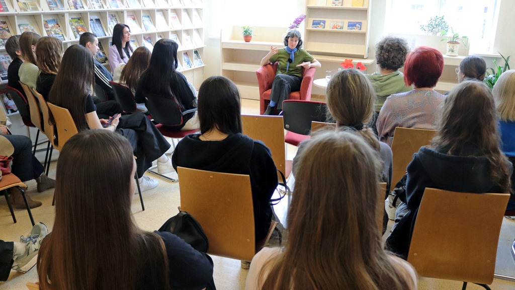 Sieviete uzrunā uzmanīgu auditoriju bibliotēkā. Viņa sēž sarkanā atzveltnes krēslā, aiz kura atrodas grāmatu plaukti, radot saistošu, izglītojošu atmosfēru.