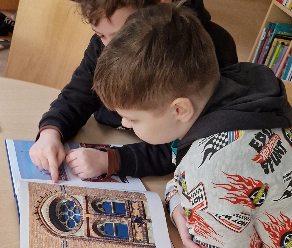Divi bērni sēž pie galda, iegrimuši grāmatā par arhitektūru. Viens bērns norāda uz attēlu, kurā redzama ķieģeļu ēka ar sarežģītiem logiem.