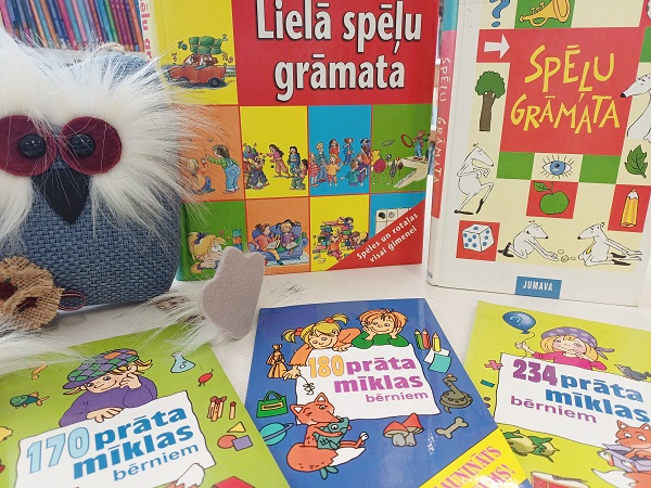 Bērnu mīklu grāmatas košās krāsās ir izliktas blakus mīlīgam zilam plīša pūces tēlam ar lielām acīm. Tonis ir rotaļīgs un aicinošs.